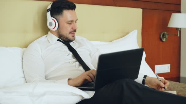 Happy businessman in headphones working at laptop computer and listening music smiling while lying in bed at hotel room. Travel, business and people concept — Stock Video