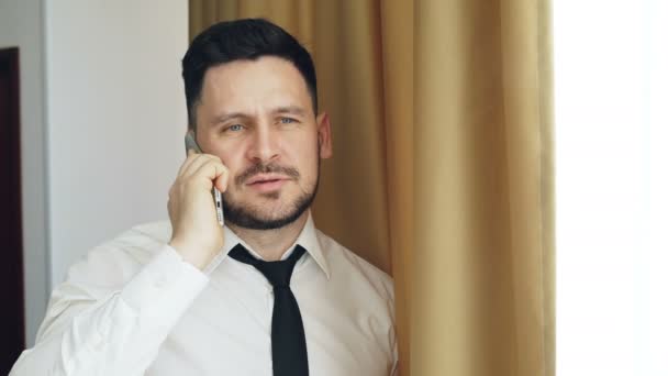 Adult bearded businessman in white shirt and tie talking on mobile phone standing near window in hotel room — Stock Video