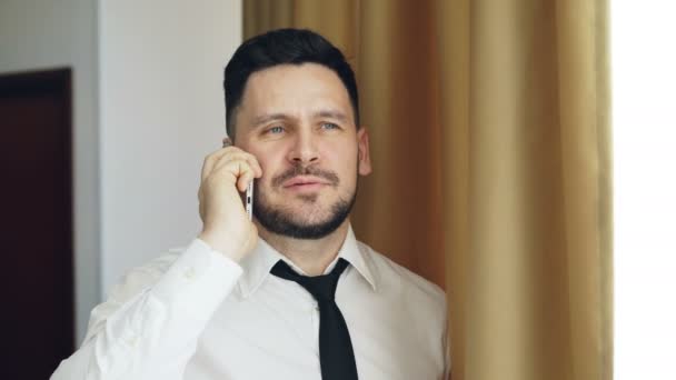 Adulto hombre de negocios sonriente en camisa blanca y corbata hablando en el teléfono móvil de pie cerca de la ventana en la habitación de hotel — Vídeos de Stock