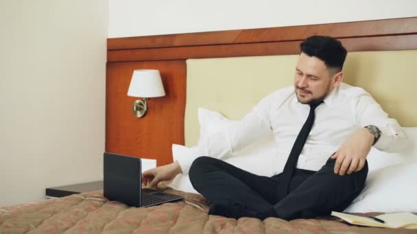 Улыбающийся бизнесмен отключил ноутбук и занялся йогой в позе лотоса, расслабившись на кровати в номере отеля. Путешествия, бизнес и люди — стоковое видео