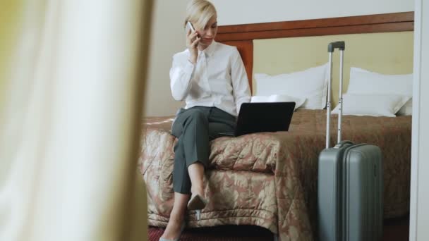 ПАН улыбающейся бизнесвумен в белой рубашке, которая пользуется ноутбуком и разговаривает по мобильному телефону, сидя на кровати в гостиничном номере. Концепция бизнеса, путешествий и людей — стоковое видео