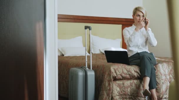 Mujer de negocios de confianza en camisa blanca usando en el ordenador portátil y hablando en el gesto del teléfono móvil mientras está sentado en la cama en la habitación de hotel. Concepto de negocios, viajes y personas — Vídeo de stock