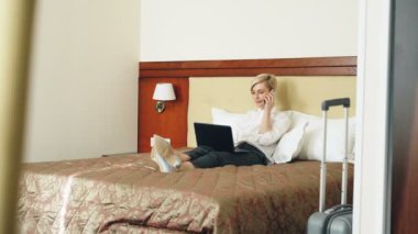 Dizüstü bilgisayarda kullanma ve otel odasında yatakta yatarken cep telefonu konuşurken beyaz gömlek gülümseyen iş kadını. İş, seyahat ve insanlar kavramı