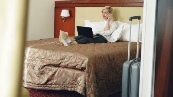 Empresária sorridente de camisa branca usando no laptop e conversando no celular enquanto estava deitada na cama no quarto de hotel. Conceito de negócios, viagens e pessoas — Vídeo de Stock