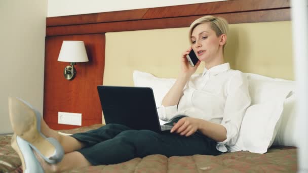 Smiling businesswoman in white shirt using on laptop and talking at mobile phone while lying on bed in hotel room. Business, travel and people concept — Stock Video