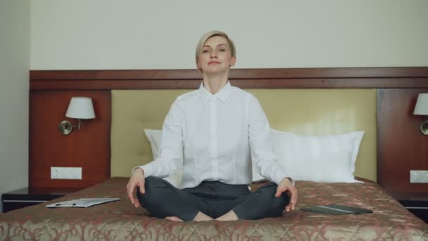 Mujer de negocios sonriente tomando posición de loto de yoga sentada relajada en la cama en la habitación de hotel. Concepto de viajes, negocios y personas — Vídeos de Stock