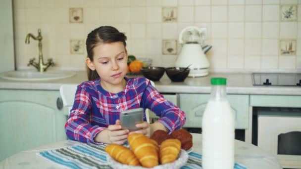 Piccola bella ragazza giocando smartphone al mattino mentre seduto a tavola in cucina. Infanzia, persone e concetto tecnologico — Video Stock