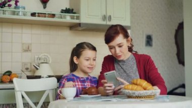 Neşeli anne ve smartphone birlikte tarama ve sabahları kahvaltı sırasında evde mutfak gülümseyen şirin kızı