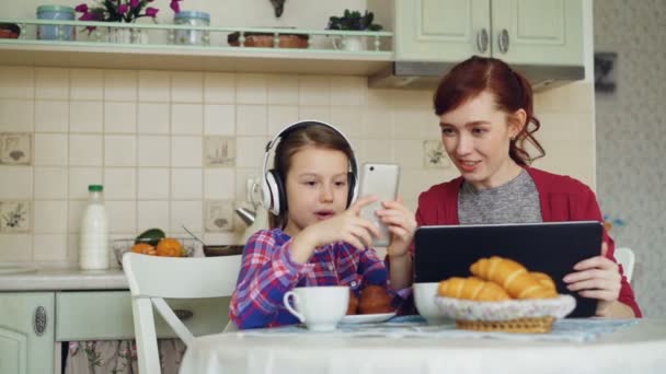 快乐的母亲和可爱的女儿早上在家里的厨房里吃早饭。小女孩玩 smarphone, 而她的妈妈浏览平板电脑 — 图库视频影像