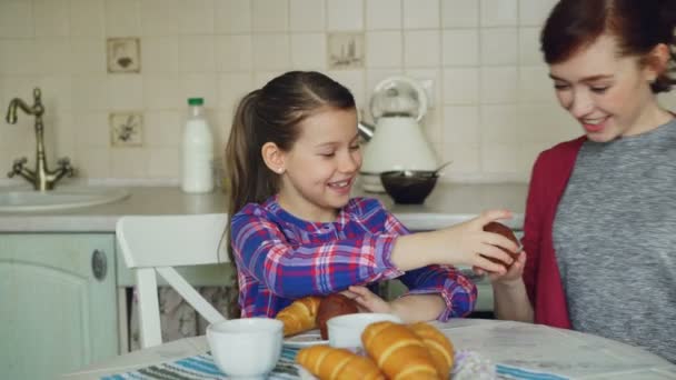 Glückliche Mutter und Tochter sitzen am Tisch und teilen Muffins und haben Spaß beim Frühstück zu Hause in der Küche am Morgen — Stockvideo