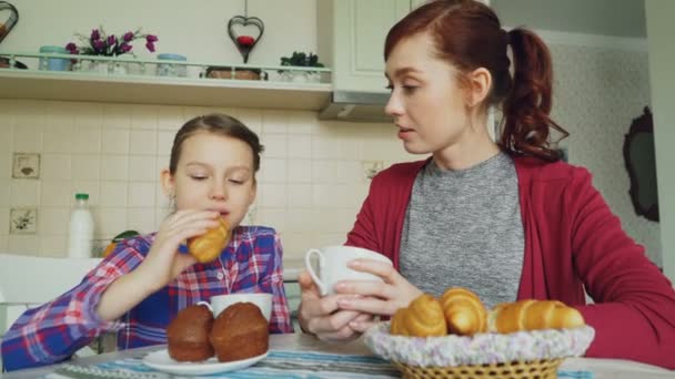 Vrolijke moeder en schattige dochter ontbijten, muffins en croissants praten op eten thuis in de moderne keuken. Familie, voedsel, huis en mensen concept — Stockvideo