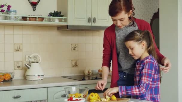 Joven madre bonita enseñando a su linda hija a cortar verduras correctamente. Niña cocinando junto con mamá cariñosa en casa en kithcen moderno — Vídeo de stock