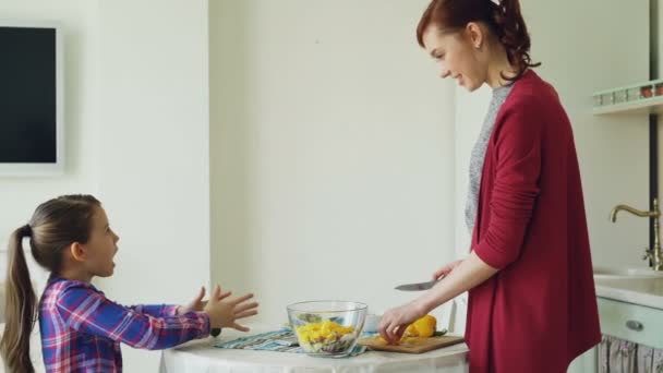 Aranyos leánya és vidám anya beszél a modern konyha főzés közben. Anya etetése lány darab növényi mosolyogva. Család, élelmiszer, home és emberek koncepció — Stock videók