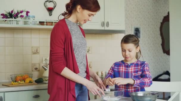Niña alegre ayudando a su madre en la cocina a cortar el huevo en un tazón y mezclando masa para galletas. Concepto de familia, comida, hogar y gente — Vídeos de Stock