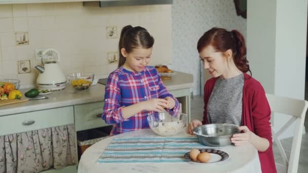 Vicces kislány segíti az anyja a konyhában, tojás, tálba vágás és keverés a tészta cookie-kat. Család, élelmiszer, home és emberek koncepció — Stock videók