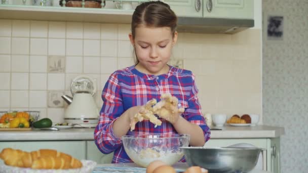 Маленька мила дівчинка готує тісто і чистить руки, посміхаючись на кухні вдома. Вона хоче пригостити свою матір печеним печивом — стокове відео