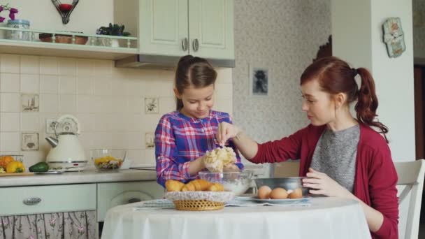 Милая девушка помогает своей матери на кухне перемешивать тесто для печенья в миску. Мама весело мажет дочь нос мукой, и они смеются. Концепция семьи, еды, дома и людей — стоковое видео