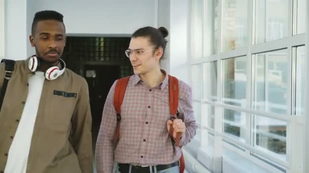 Dos guapos estudiantes masculinos caminando en un largo corredor de cristal blanco en Universit conociendo a sus compañeras de grupo diciendo hola de pie y hablando positivamente — Vídeos de Stock