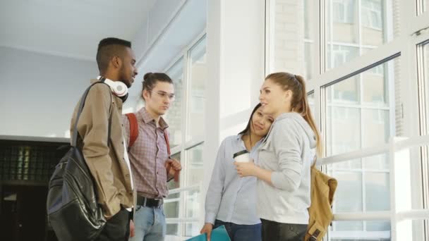 Quatre étudiants multi-ethniques sont debout dans une grande salle blanche spacieuse à l'université parler entre eux de manière positive. Ils sourient positivement et rient — Video