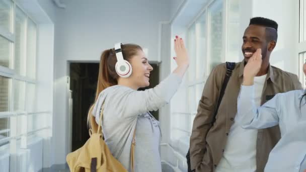 Dolly vue de quatre étudiants masculins et féminins d'ethnie différente debout près de la fenêtre dans un large couloir blanc discutant étude souriant et riant — Video