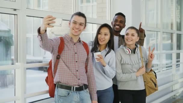 Un groupe de quatre étudiants multiethniques positifs, hommes et femmes, se tient dans un large couloir. Hipster intestin tient smartphone faisant selfie de tous — Video