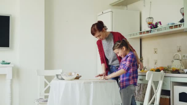 Joven madre enseñando a su pequeña hija linda rodando masa mientras cocinan juntos en la cocina en casa en las vacaciones. Concepto de familia, comida y personas — Vídeos de Stock