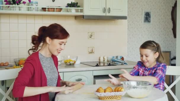 Молодая мать и милая дочь готовят вместе, разговаривая на кухне в выходные. Девочка хлопает в ладоши мукой и смеется. Концепция семьи, еды, дома и людей — стоковое видео