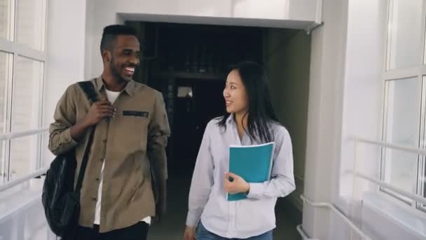 Fiatal vonzó afrikai férfi és ázsiai női hallgatók valamit élénk és intett, hogy egy másik pár állandó magas ablak közelében elhaladó megvitatni folyosón haladunk — Stock videók