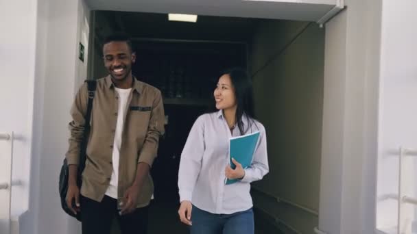 Молодой симпатичный афро-студент и его девушка ходят по коридору колледжа и что-то обсуждают. Они встречают друзей, стоящих у окна и размахивающих ими — стоковое видео