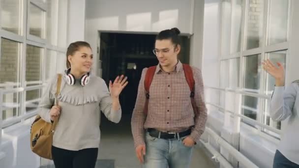 Två kvinnliga och manliga studenter vandrar i Universitetshuset diskutera något. De möter vänner stå nära fönstret killen vifta med dem — Stockvideo