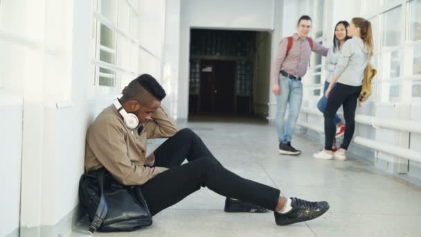 Upprörd allvarliga afroamerikansk manlig student sitter på golvet i universitet korridor ute i olika sidor medan hans groupmates står kommunicera — Stockvideo