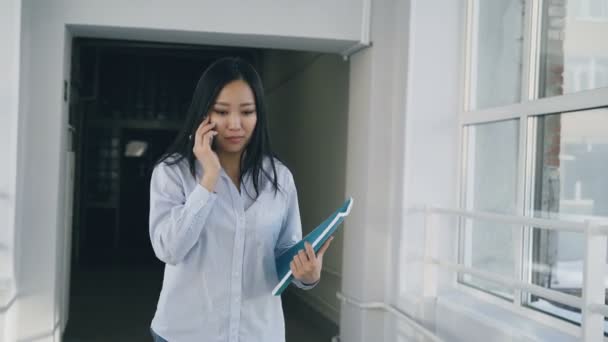 Dolly skott av asiatiska kvinnlig student promenader i brett universitet korridoren pratar telefon förbi andra multietniska studenter — Stockvideo