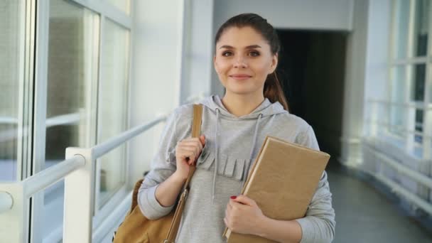 Portrait of young beautiful caucasian confident female student standing in white glassy corridor smiling positively holding textbook looking at camera — Stock Video