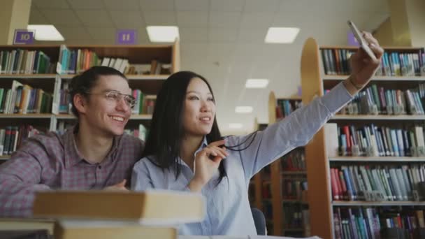 Молодая пара подростков, сидящих за столом в библиотеке в окружении книг, готовящихся к экзамену. Девушка держит своего умника и делает селфи со своим одноклассником — стоковое видео
