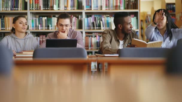 Multietnická parta studentů a umístění v knihovně s knihami a laptop na stole, příprava k vyšetření společně usmívá a směje se — Stock video