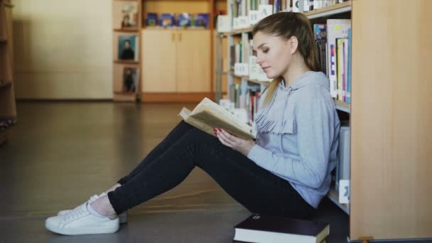 Joven hermosa mujer caucásica estudiante en ropa casual está sentado en el suelo en una gran biblioteca espaciosa cerca de librería lectura interesante libro grande en serio — Vídeos de Stock