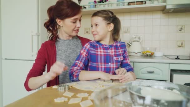 Vrolijke moeder en schattig dochtertje praten en het maken van cookies samen met bakkerij formulieren bijsnijden deeg zittend in de moderne keuken thuis. Familie, eten en mensen concept — Stockvideo