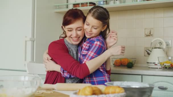 Alegre madre cocinando en la cocina mientras linda hija viene y abrazando a mamá por la mañana. Concepto de familia, comida, hogar y gente — Vídeos de Stock