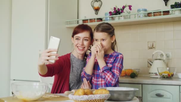 Smiling mother together with funny daughter taking selfie photo with smartphone camera making silly face while cooking at home in kitchen. Family, cook, and people concept — Stock Video