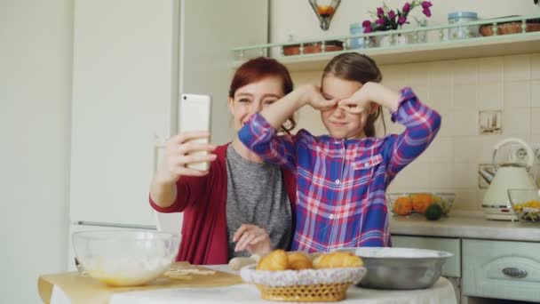 Mère souriante avec fille drôle prenant des photos selfie avec appareil photo smartphone faire visage stupide tout en cuisinant à la maison dans la cuisine. Concept de famille, cuisine et personnes — Video