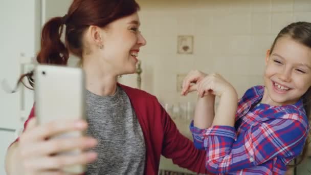 Sonriente madre junto con divertida hija linda juguetona haciendo foto selfie con cámara de teléfono inteligente en casa en la cocina. Concepto de familia, cocinero y gente — Vídeos de Stock