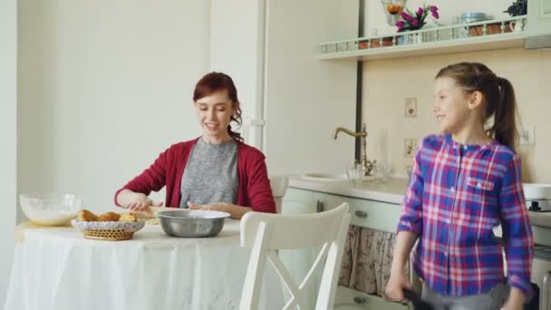 Joven madre rodando masa mientras cocina y su divertida hija caminando y cantando con peine bailando en la cocina el fin de semana. Concepto de familia, comida, hogar y gente — Vídeos de Stock