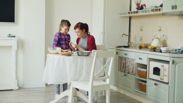 Madre sonriente y linda hija haciendo galletas juntos usando masa mientras están sentados en la cocina moderna en casa. Concepto de familia, comida y personas — Vídeo de stock