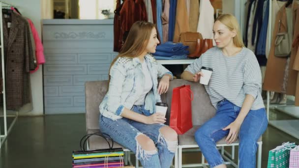 Dos chicas encantadoras sentadas en la tienda de ropa con café, hablando y riendo después de ir de compras. Bonita boutique con coloridas prendas, bolsos y zapatos en el fondo — Vídeos de Stock