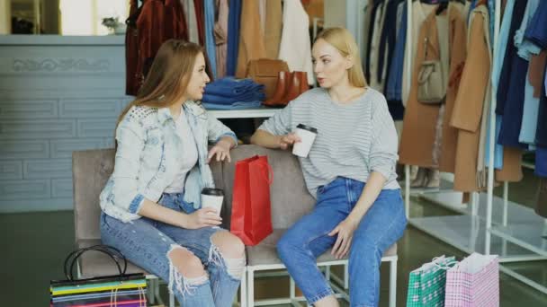 Twee jonge vrouwen zitten op stoelen leer bedekte in kleding boetiek en chatten na het winkelen. Mooie moderne womens slaan met veel kleding in achtergrond — Stockvideo