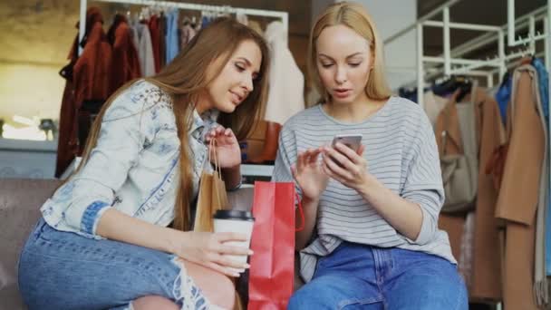 Alegre mujeres jóvenes charlando y utilizando un teléfono inteligente moderno mientras se sienta en la tienda de ropa agradable. Sonríen y hacen gestos con entusiasmo. . — Vídeo de stock