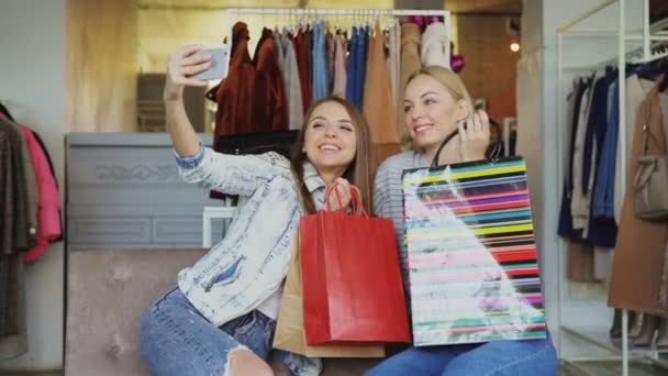 Las chicas lindas están haciendo selfie con bolsas de papel coloridas en la boutique de ropa de mujer usando un teléfono inteligente, y luego viendo fotos juntas. Sonríen y ríen descuidadamente . — Vídeos de Stock