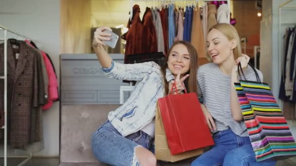 Amigas atraentes estão usando o smartphone para fazer selfie enquanto estão sentadas na loja de roupas com sacos de papel coloridos. Eles estão sorrindo, posando, rindo, gesticulando . — Vídeo de Stock