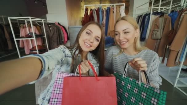 Punto de vista plano de dos chicas descuidadas atractivas haciendo selfie con bolsas de papel en la tienda de ropa de las mujeres. Los amigos están posando, riendo y charlando felizmente — Vídeos de Stock