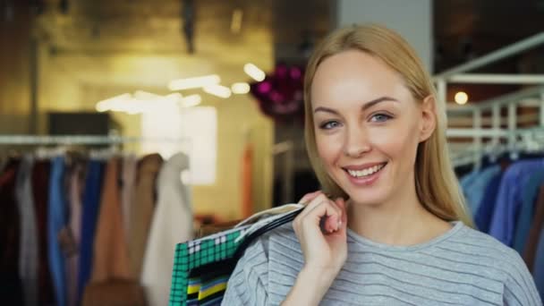 Retrato de cerca de una chica rubia de pie con bolsas de papel en la boutique de ropa, riendo y sonriendo felizmente y mirando a la cámara. Ropa con estilo está en el fondo . — Vídeo de stock